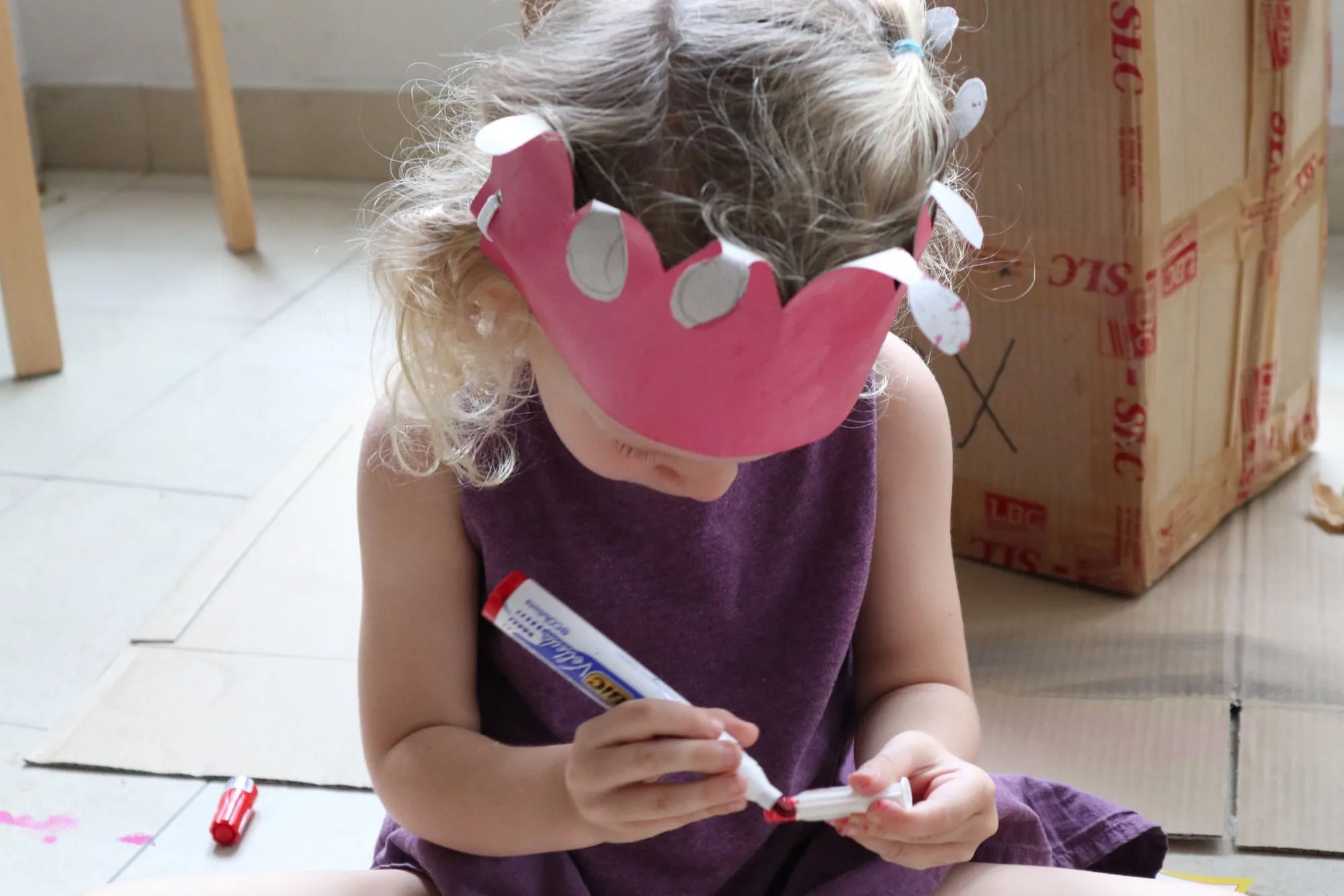 A little girl in a paper crown colouring with a marker on cardboard